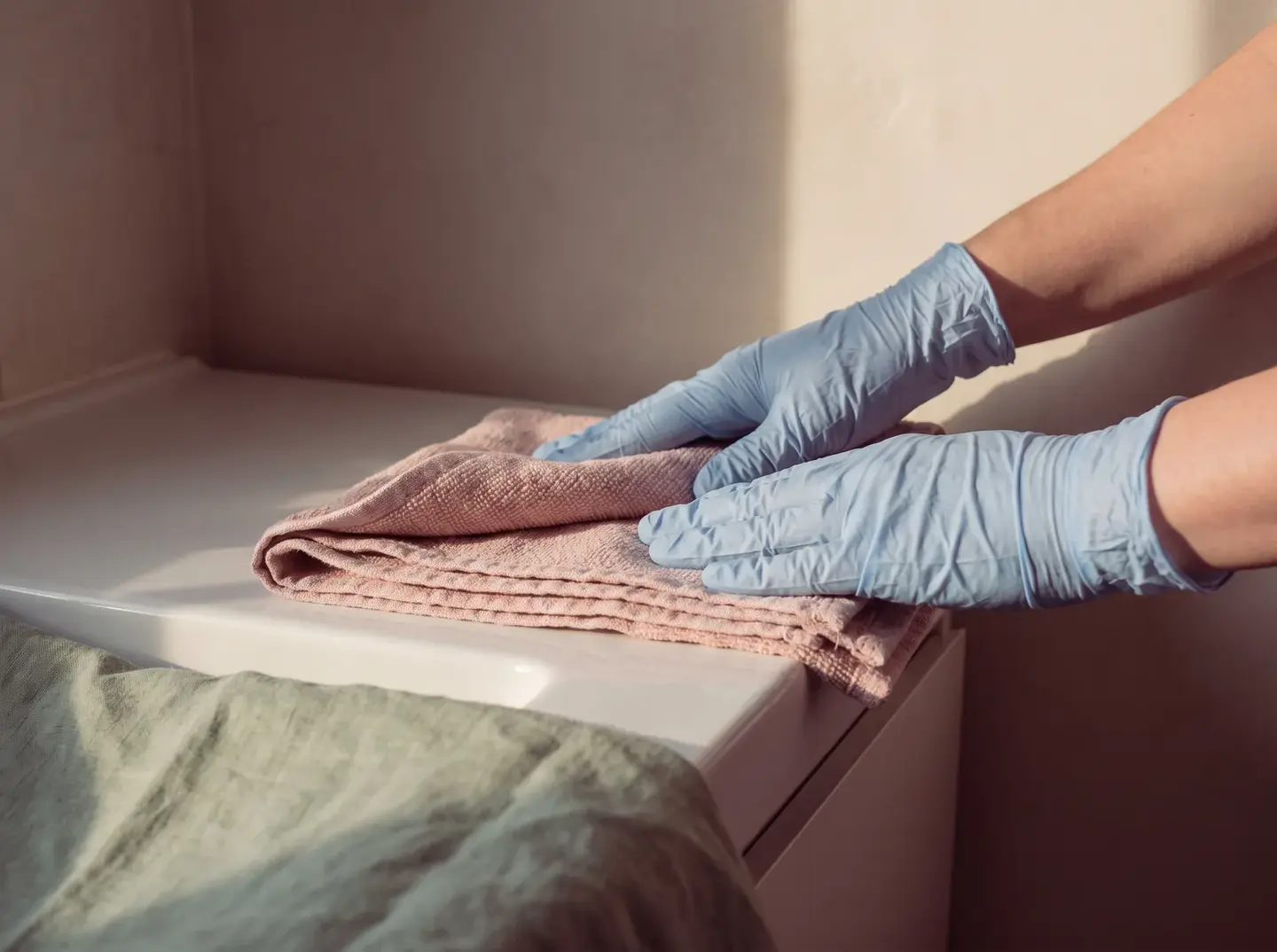 Manos con guantes claros doblan despacio una toalla rosa pálido bajo una luz lateral cálida